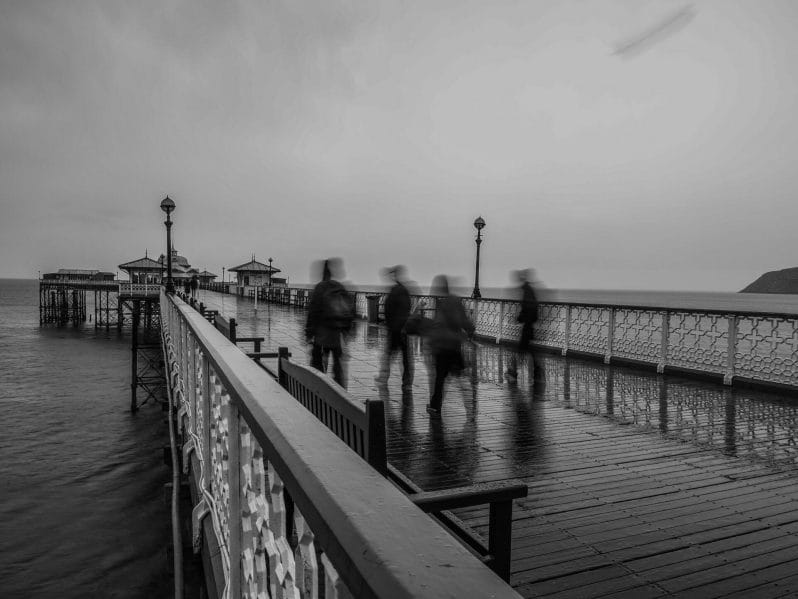 Black and White Photo of four photographers (blurred) being captured moving while walking along the Llandudno Pier in North Wales