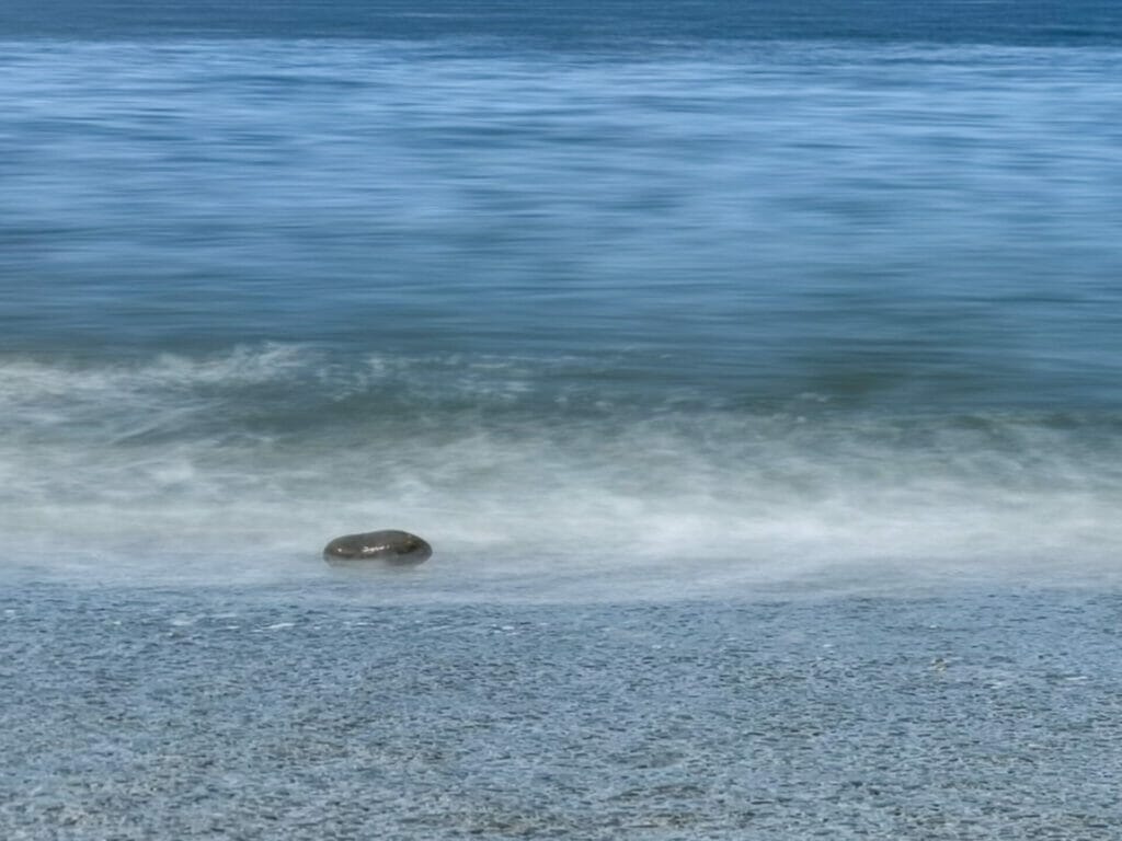 A blurred photo of sand, sea and blue sky in Llandudno showing a rock with small waves washing over it. 