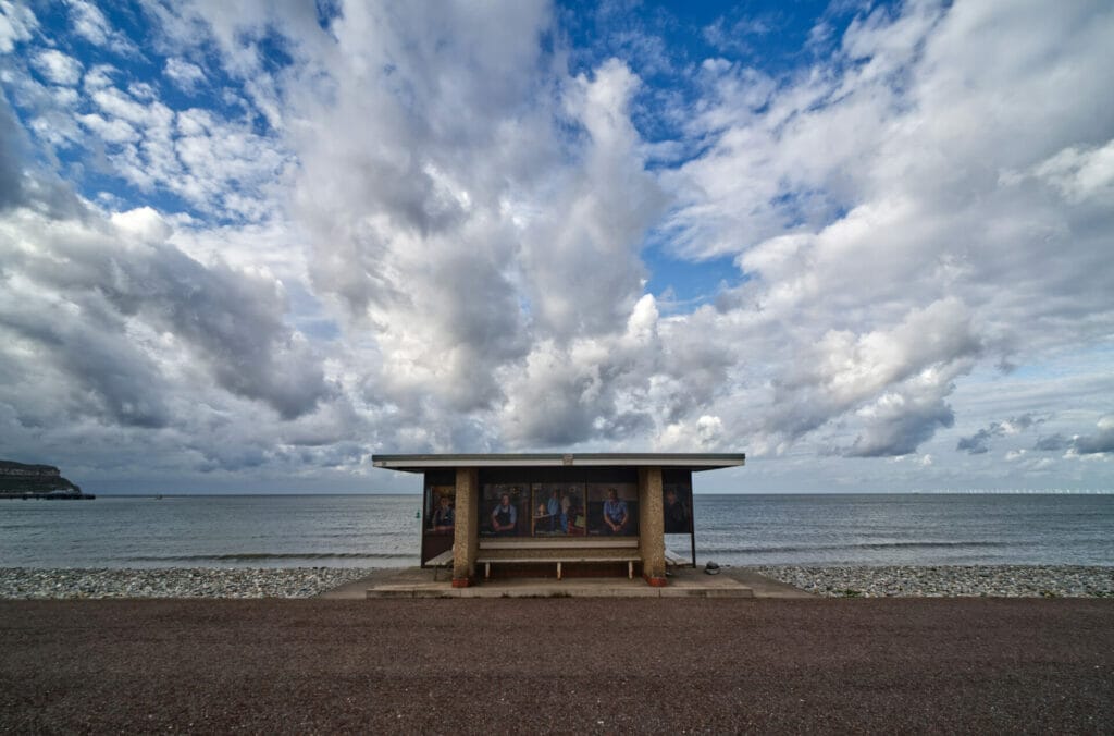 Photograph of a dramatic blue sky and white clouds above a shelter on the Llandudno prom in North Wales with photographic images of people working in hospitality which is part of a project. Photo taken on a Taken on a Welshot Photographic Workshops and Events