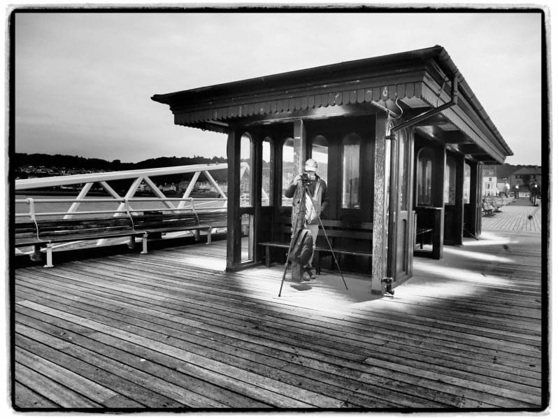 Black and White Photo of the shelter on Beaumaris Pier on Anglesey with a photographer taking a photo on a Welshot Photographic Workshop
