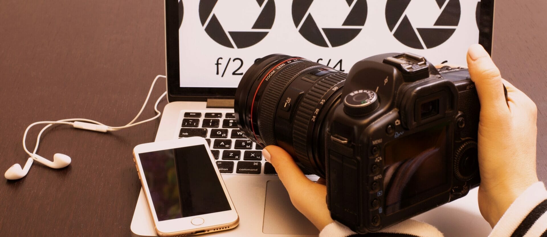 I Want To Learn How To Use My Camera - Photo of a person holding a camera, looking at the back of it while it sits on an open laptop with a phone next to it.