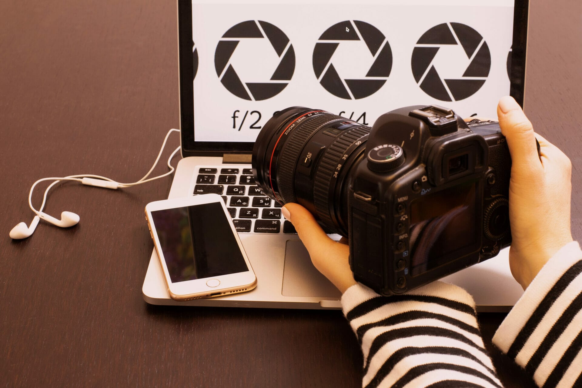 I Want To Learn How To Use My Camera - Photo of a person holding a camera, looking at the back of it while it sits on an open laptop with a phone next to it.