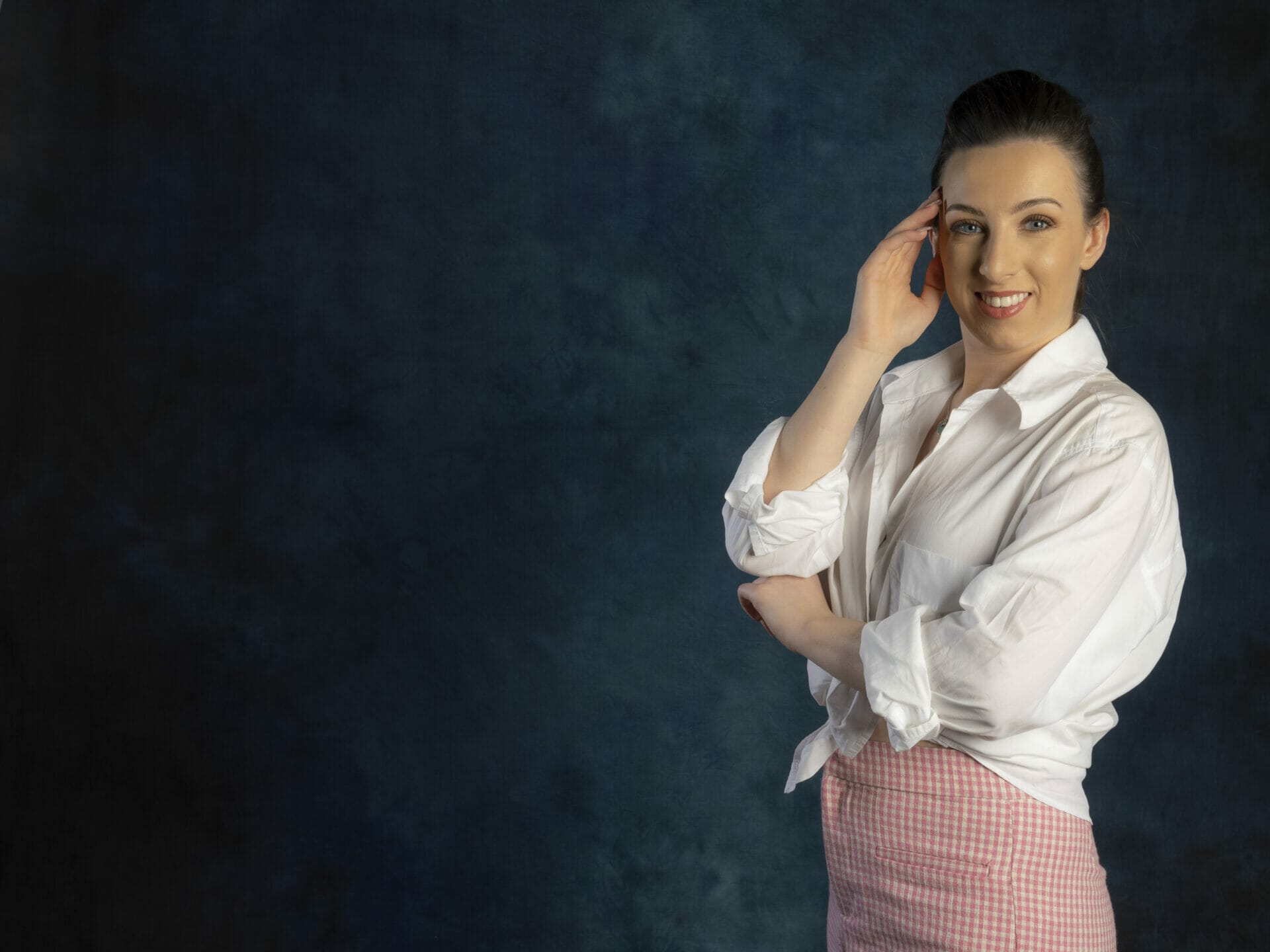 Image of a young lady wearing a white shirt and pink and white check 1960's trousers - with a dark blue background - taken on a Friday Night is Studio Night Portfolio Building Session with the Welshot Photographic Academy in Llandudno, North Wales