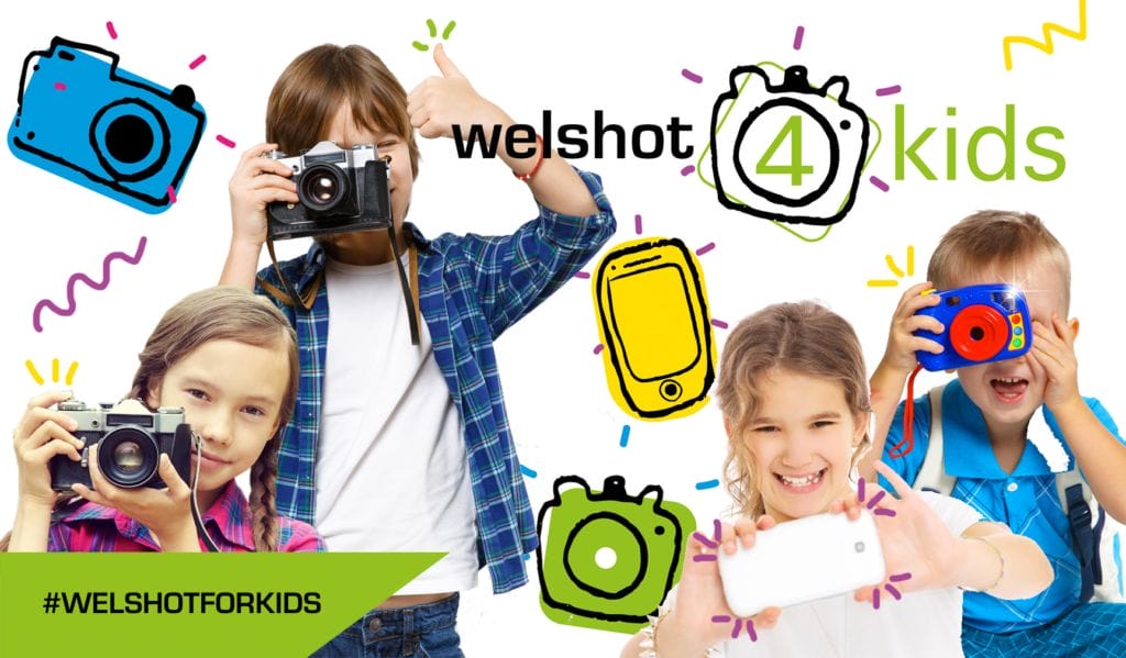 A colour photo showing four children of varying ages playing with cameras. there are also some cartoonie graphics (cameras) interspersed around the children with the wording Welshot for (showing the numeral 4 as the for)) Kids